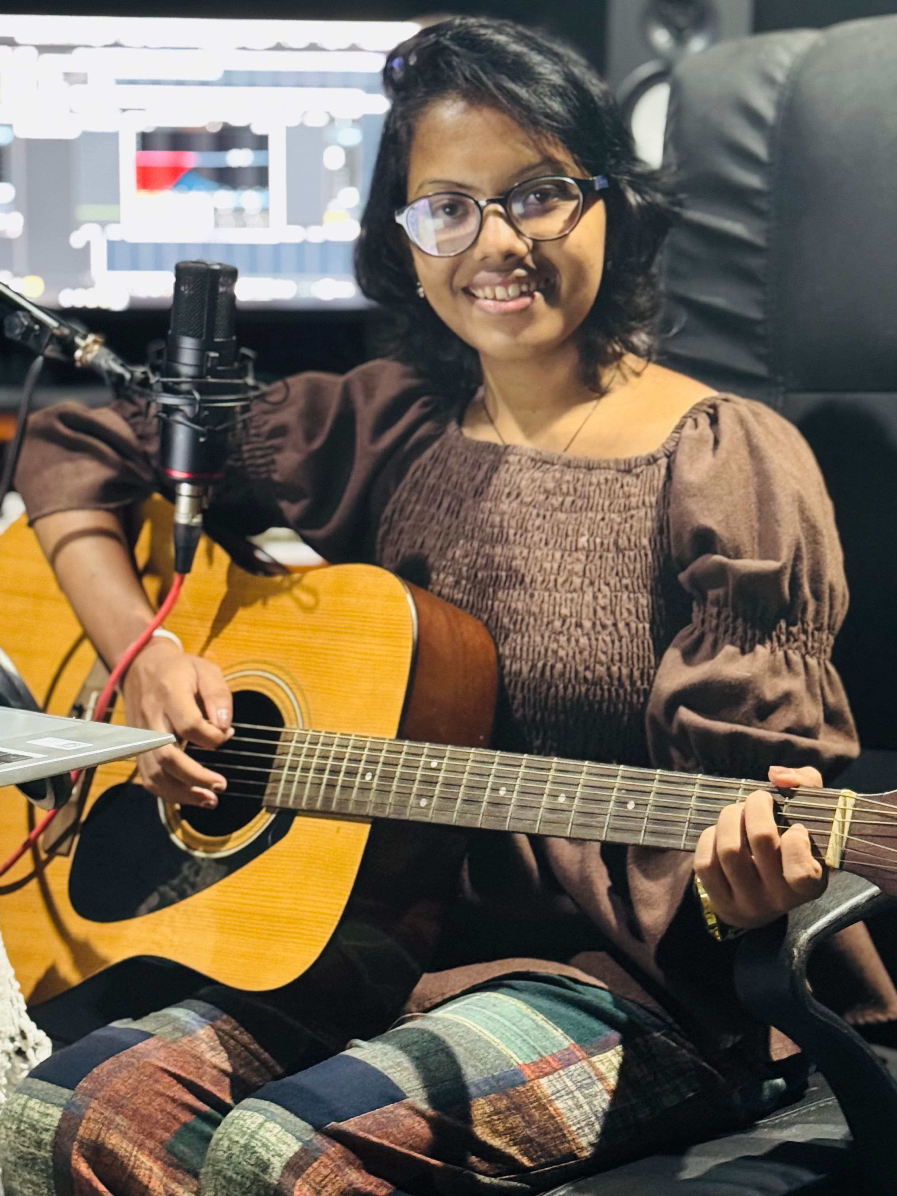 Artist seated with an acoustic guitar beside a microphone inside the Tastic Wavs studio.