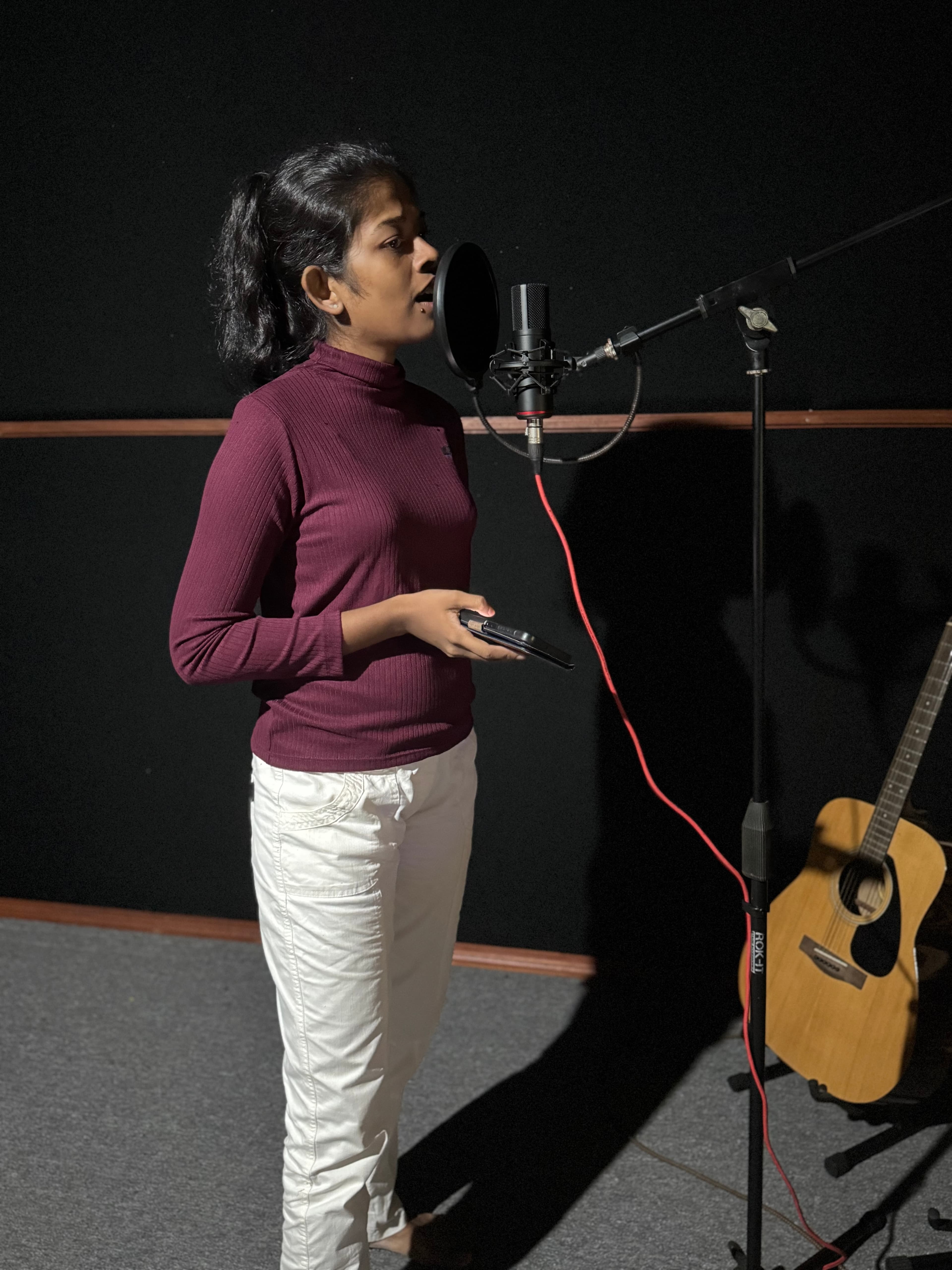 Singer performing into a studio microphone while holding a phone for lyrics or notes.