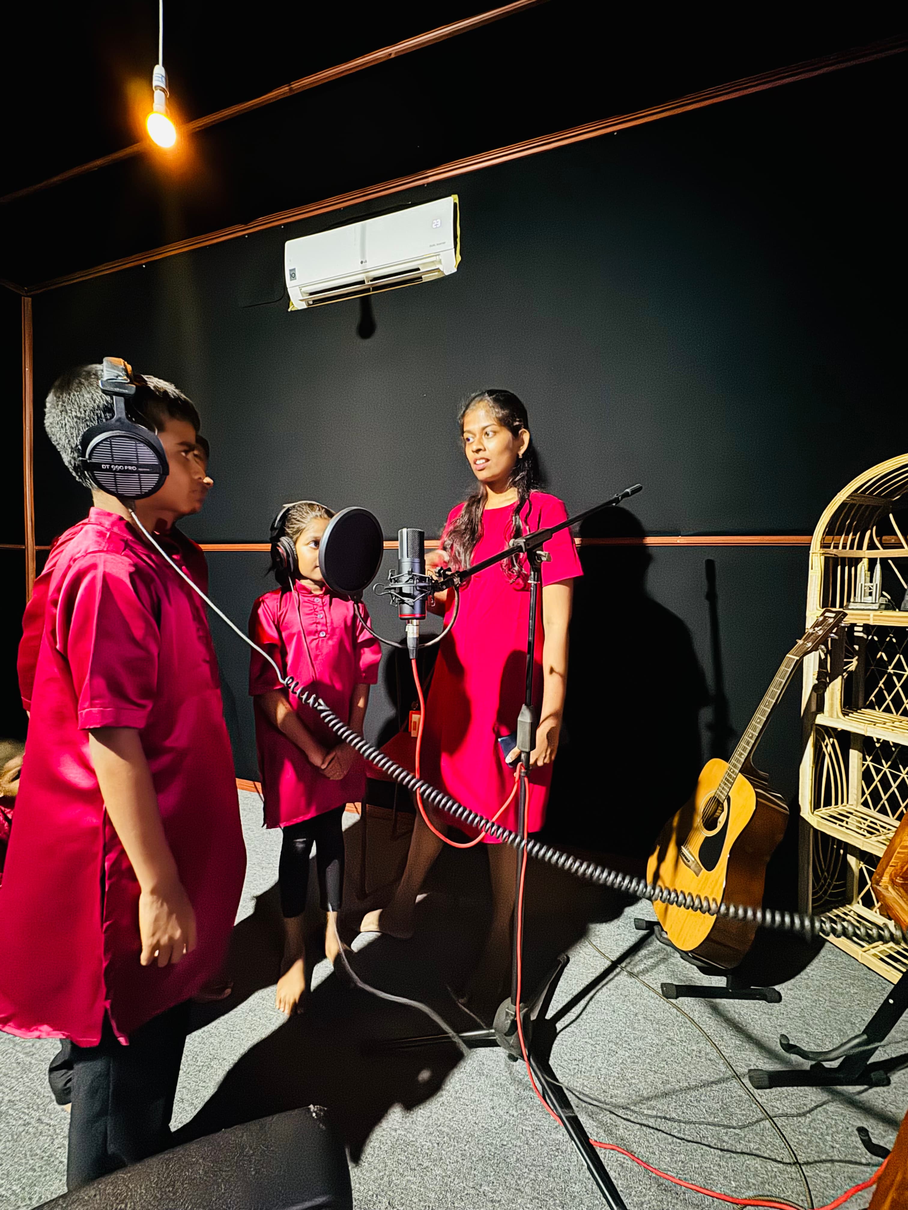 Three young performers gathered around a studio microphone during a recording session.
